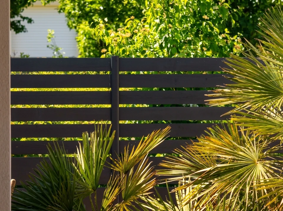 Jardin extérieur ensoleillé avec clôture moderne à lames horizontales en aluminium laqué anthracite, fixées sur poteaux carrés noirs