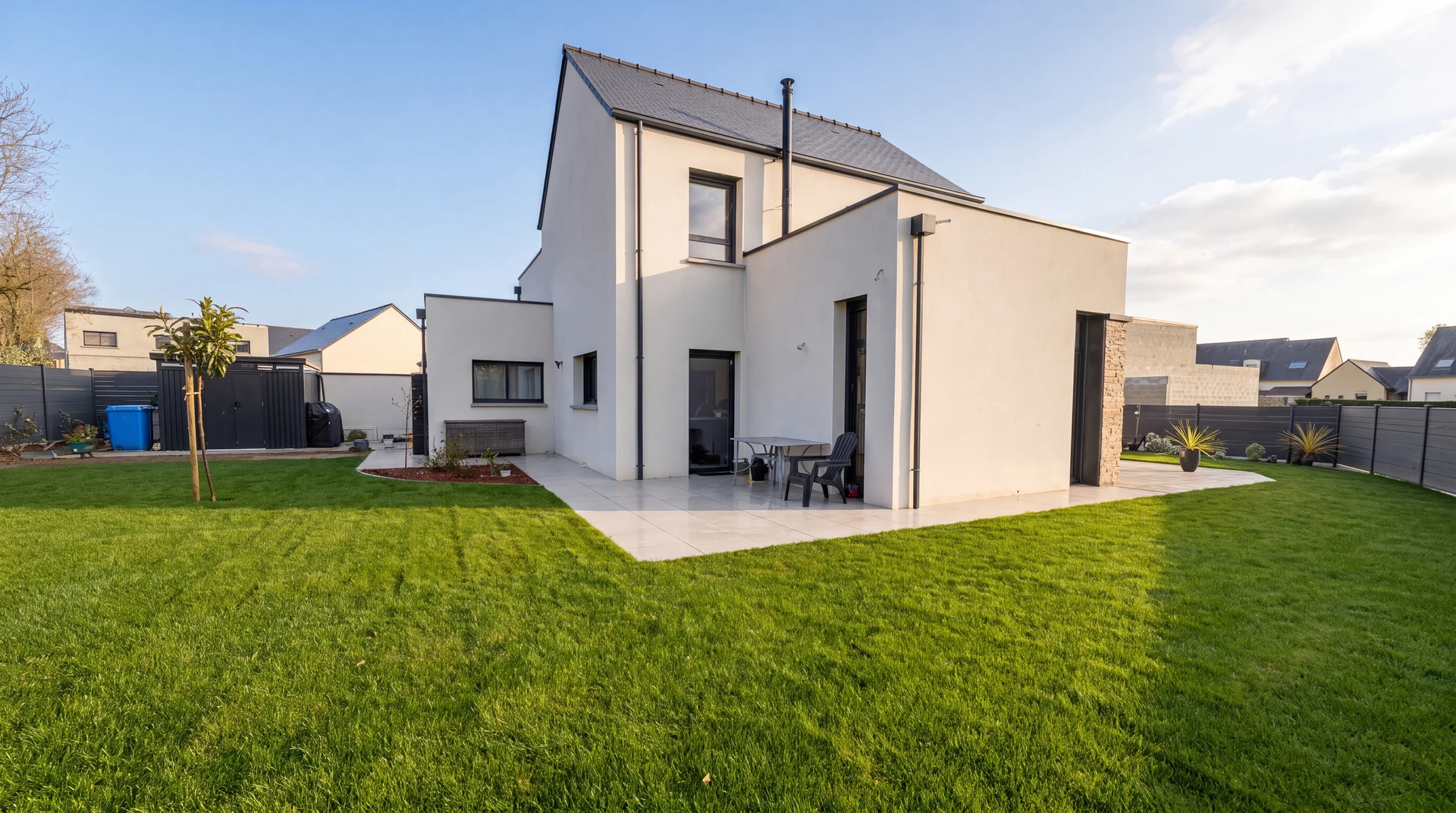 Jardin résidentiel en friche avec pelouse haute non entretenue, maison moderne à deux niveaux avec extension single-story, terrasse bois composite, clôtures périmétriques en panneaux bois horizontal et vertical
